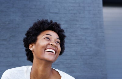 Fotografía de una mujer afrodescendiente, de pelo corto, sonriendo con la mirada hacia arriba. Detrás un fondo de ladrillo gris