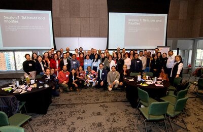Fotografía de grupo durante la Consulta Regional para la Priorización de la Investigación sobre TCIM en las Américas, celebrada los días 11 y 12 de junio de 2025 en São Paulo (Brasil). Aproximadamente 50 participantes posan sonrientes en el centro de una sala con mesas y materiales de trabajo. Al fondo, dos pantallas muestran el orden del día de la sesión "Temas y Prioridades de la MT".