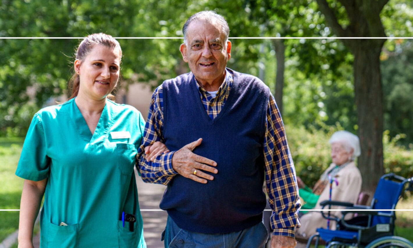 older man and nurse walking in wooded area with an older women in a wheelchair in the background